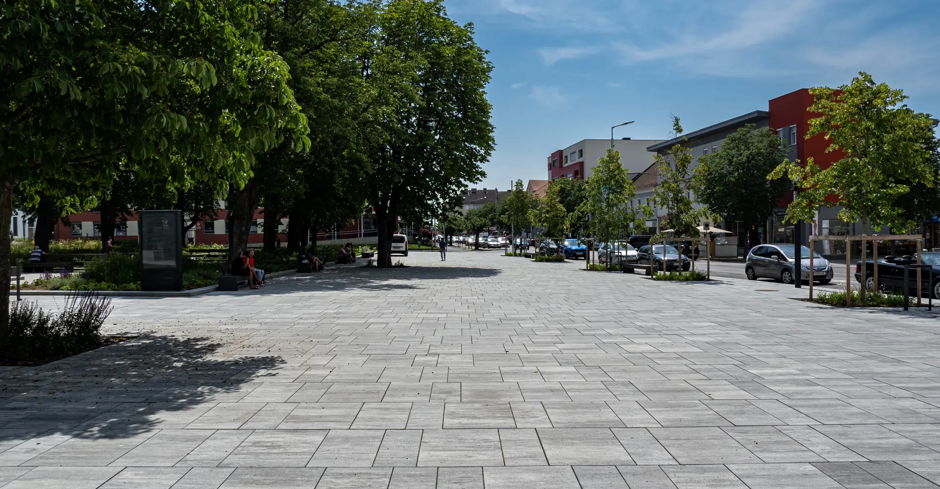 Wegefläche im Stadtgarten Oberwart mit Magnum Platten granitgrau-schattiert, gesäumt von Bäumen und Beeten, Friedl Steinwerke.