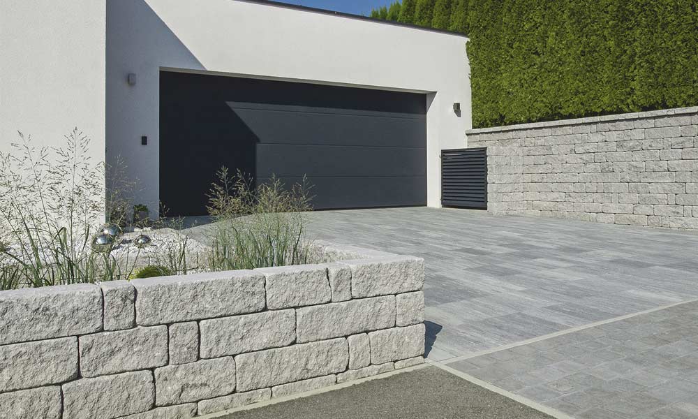 Mauerstein Gutshof MB16 bossiert grau als Begrenzung und mit Mauerbreite 24 cm (MB24) und Abdeckplatte Gutshof 3-seitig gespalten in Grau als Mauer im Hintergrund Mauerstein Gutshof MB16 bossiert grau als Begrenzung und mit Mauerbreite 24 cm (MB24) und Abdeckplatte Gutshof 3-seitig gespalten in Grau als Mauer im Hintergrund