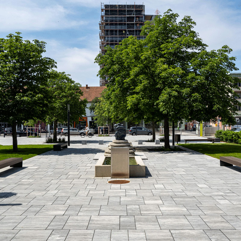 Stadtgarten Oberwart mit Brunnen und heller Platzfläche unter großen Bäumen; Magnum granitgrau-schattiert, Friedl Steinwerke.